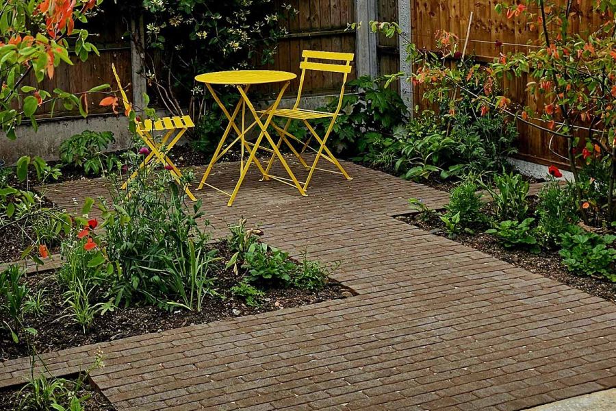 Yellow metal furniture sits on an Aldridge clay paver patio arranged in a J-shaped pattern, surrounded by sunken flowerbeds.