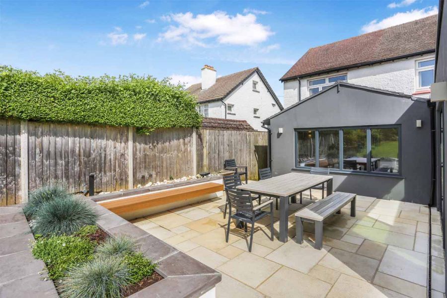 Grey extension sits next to antique cream sandstone patio used for dining area with table and chairs, raised flowerbeds and built-in bench surround.