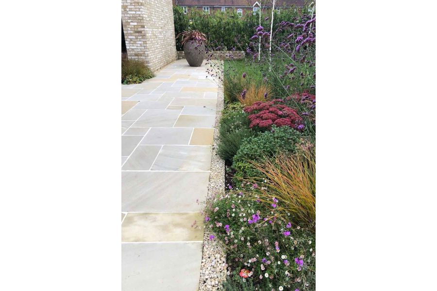 A line of pebbles runs either side of flowerbed and antique cream sandstone paving with plant pot sitting on the patio in the distance.