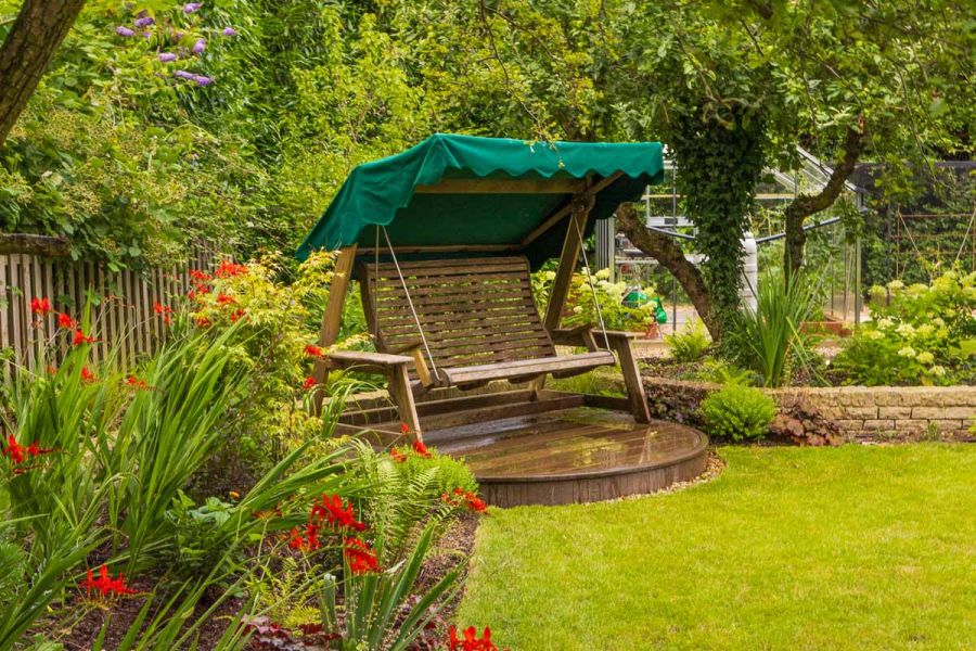 Wooden sitting swing sits in private area on antique oak millboard circle decking area surrounded by planting and large trees.