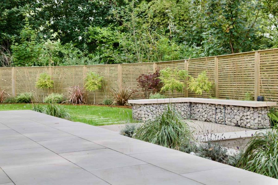 Gabion basket style bench sat on clay paver area with ash beige porcelain patio and lawn enclosed by batten fencing.