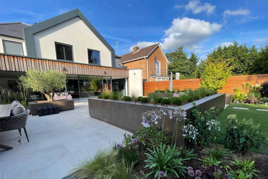 Patio of ash beige porcelain paving with views of back of house with large glass doors, raised flowerbeds create seating area.