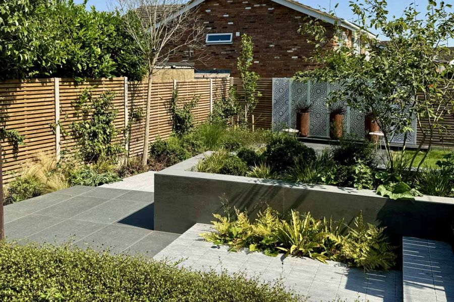 Hedges, trees and raised flowerbeds surround black basalt porcelain paving area that leads to decking and garden feature with corten panels.