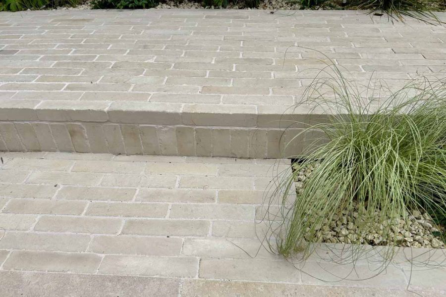 Two level patio using beige sandstone patio bricks as pavers and for cladding the step up, grassy foliage on the right.