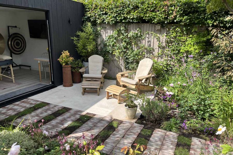 Two sunloungers sit on limestone patio, with bexhill clay paver path, outside a garden room with massage table, surrounded by planting.