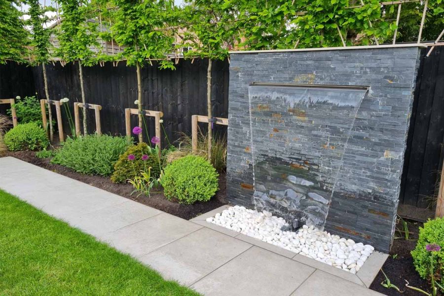 Water pours from black slate cladding waterfall feature into white pebbles with porcelain path running past.