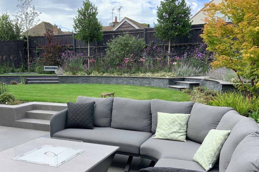 Curved sofa around a firepit, set in front of a low retaining wall that leads to a raised lawn, leading further to Black Slate External Cladding.