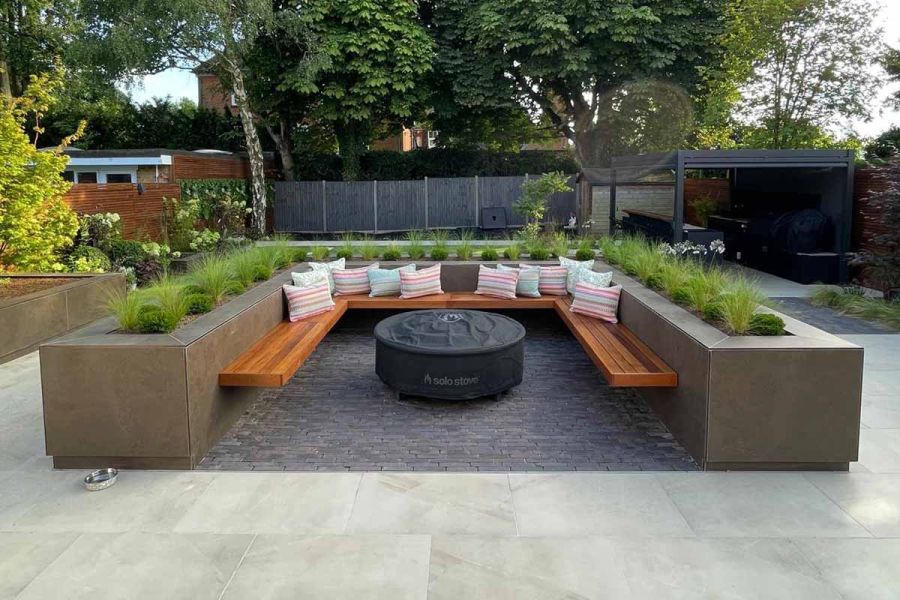 Patio with raised flowerbeds used as walls to contain floating bench and firepit, sitting on lugano clay pavers. Pergola in distance.