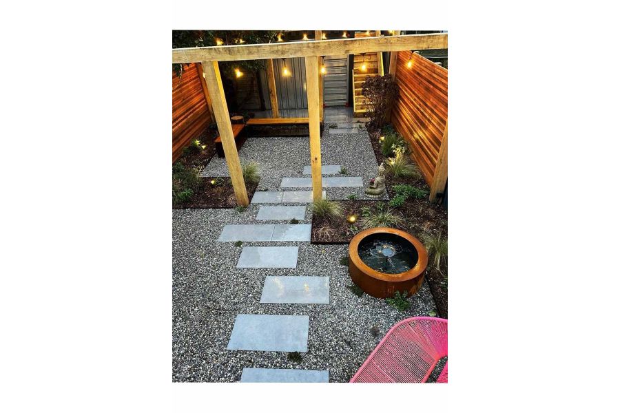Steel Corten water feature sits next to stepping stones made from steel grey porcelain paving, running under pergola with string lights at night.