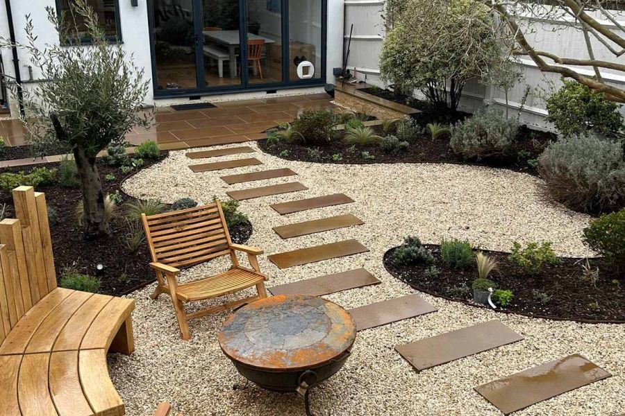 Patio of Buff sawn sandstone paving leads from french doors into pebbled area with sandstone stepping stones, flowerbeds and seating surround.