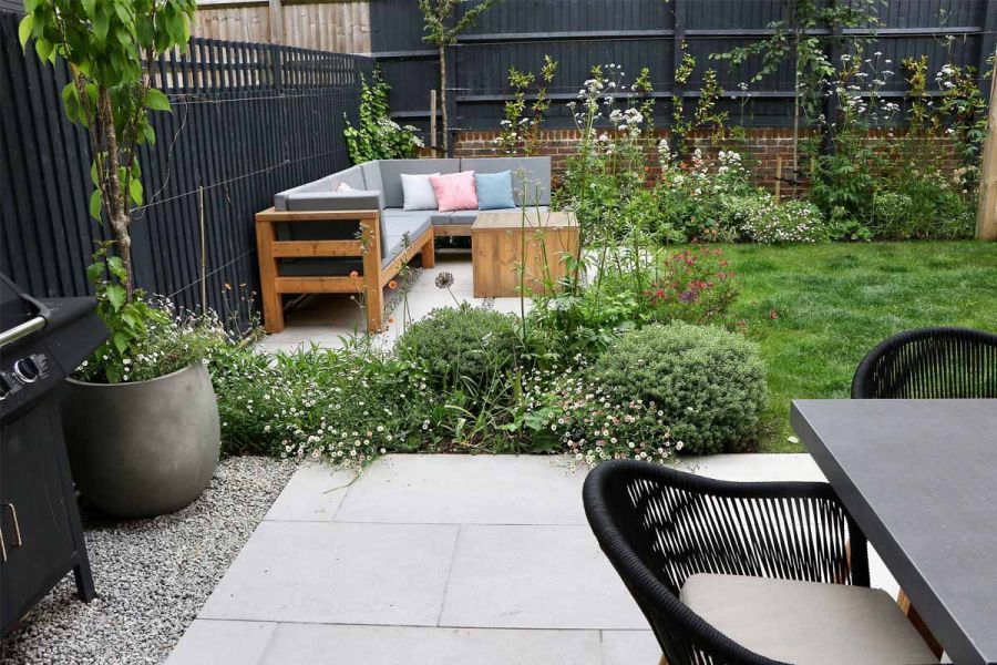 Florence Beige porcelain paving used as patio for dining set with bbq on gravel path, lawn and outdoor sofa in background.