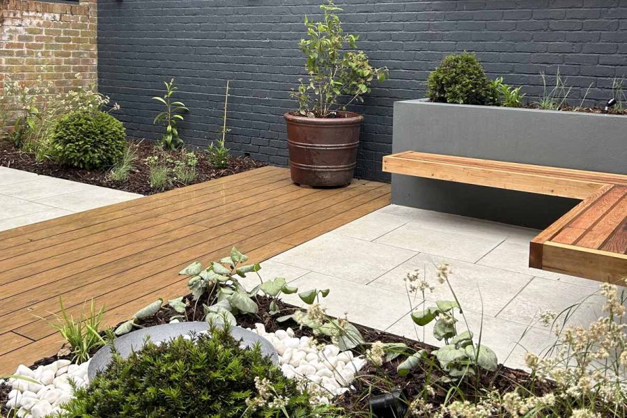 Warm teak brushed composite decking is bordered by porcelain paving, with a floating bench and lush planting around it.