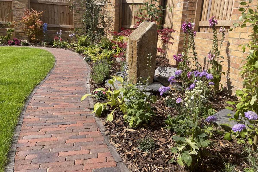 Winding path of antique red clay pavers leads through lawn and flowerbeds with purples and reds, against brick walls and fencing.