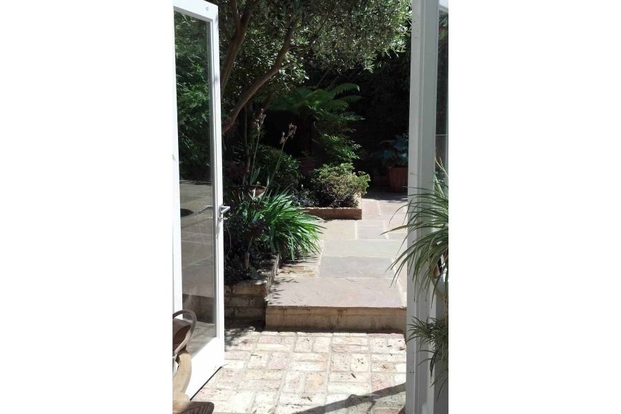 View through open french doors to brick pavers with step up to Tumbled Raj Green sandstone paving and beds with shrubs and trees.