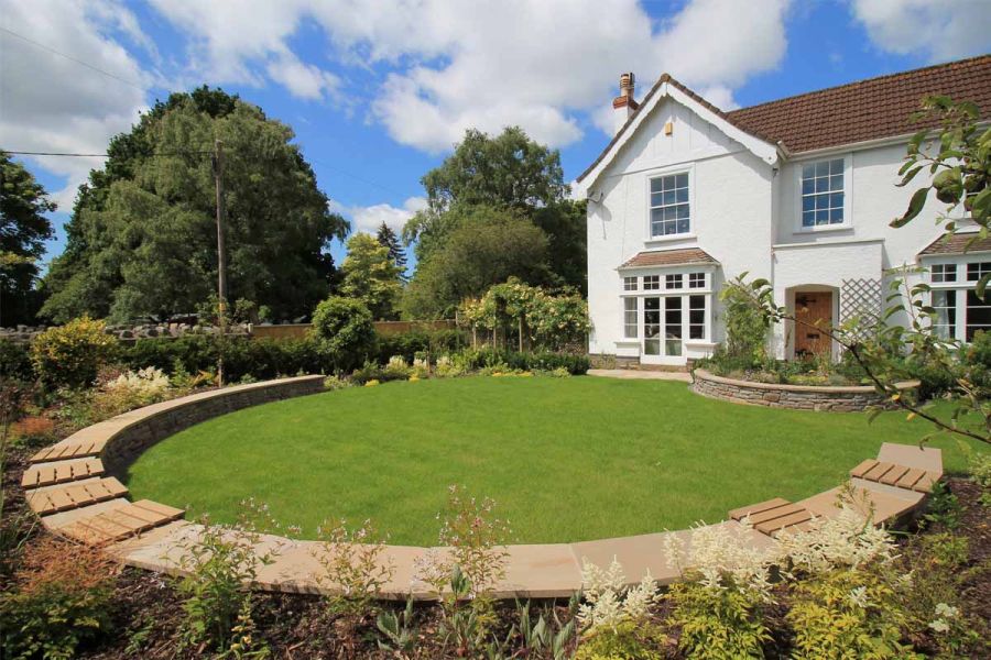 Curved, built-in bench using sandstone and warm teak composite battens surrounds lawn at the back of white painted property.