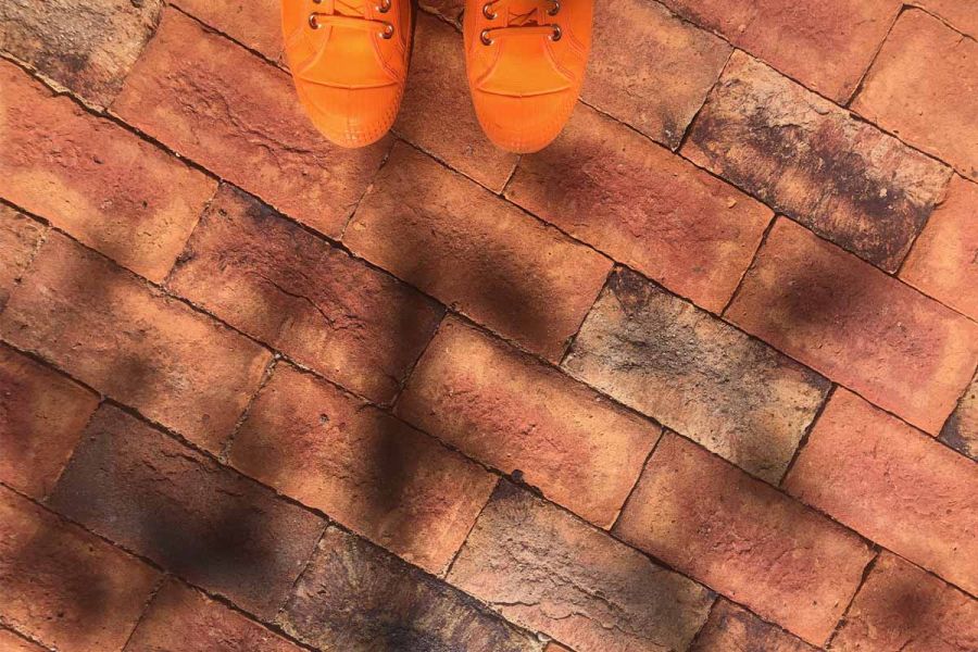 A pair of bright orange trainers stand on cotswold clay paver bricks showcasing red and dark tones with shadows casting.