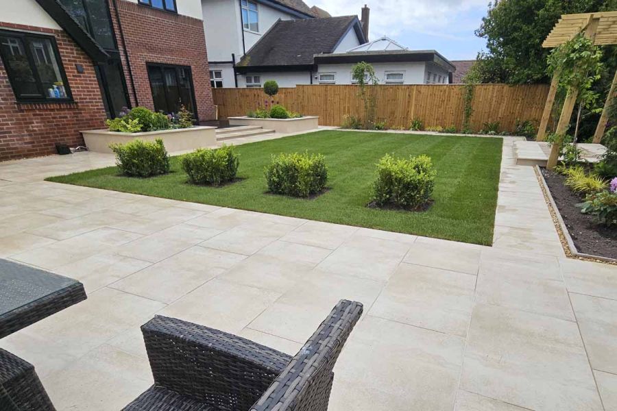 Rectangle, well-manicured lawn with 4 neatly planted bushes sits in the middle of cream porcelain paving patio.
