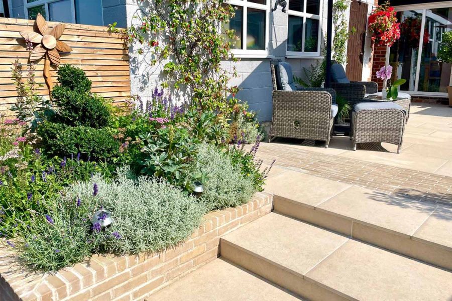 Steps made from porcelain lead up to patio indented with Westminster clay pavers, flowerbed sits next to rattan garden furniture.