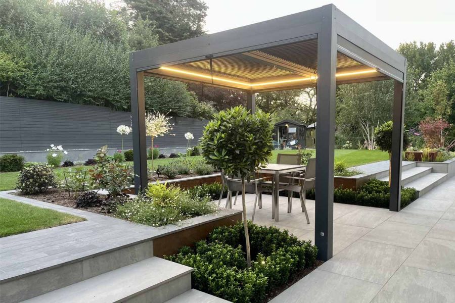 Lit up proteus pergola sits on porcelain paving with lawn and flowerbeds surrounding, enclosed by dark ash composite battens.