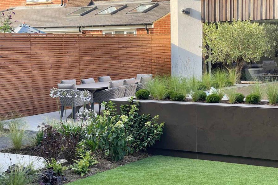 Raised flowerbeds filled with grasses and hedges, clad with dark mocha porcelain wall cladding with dining table in the distance.