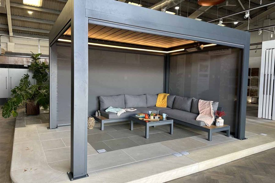 Deluxe Anthracite Grey Metal pergola with motorised blinds closed on 2 sides and LED lights on, in showroom setting.