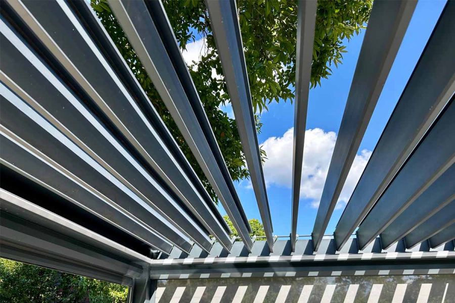 Close-up of the Deluxe Anthracite Grey 3x3 metal Pergola's open louvres, framing a backdrop of trees and blue skies.