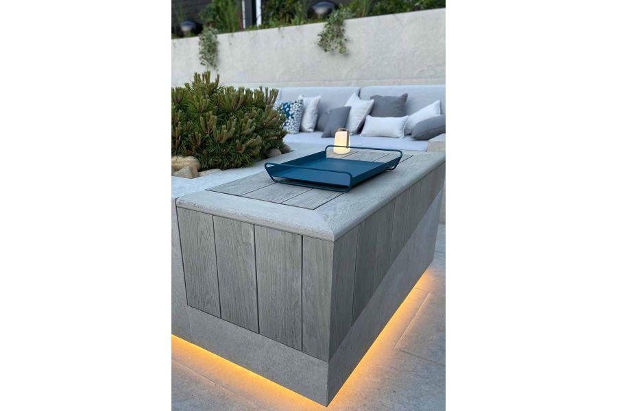 Close up of raised wall clad with smoked oak millboard with lighting underneath and drinks tray on top, built in sofa in distance.