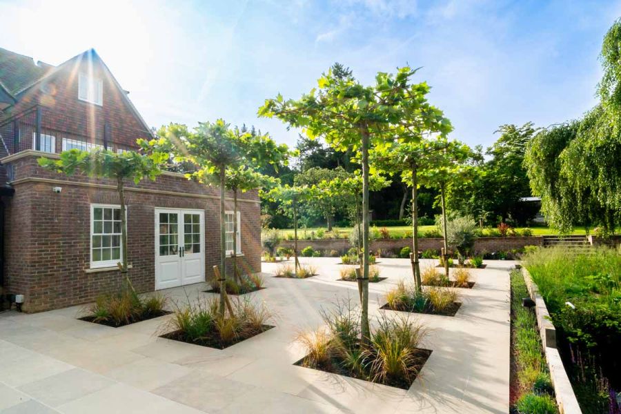 Sunshine beams onto heath sawn sandstone paving from pointed roof of large house, trees planted inbetween the patio paving.