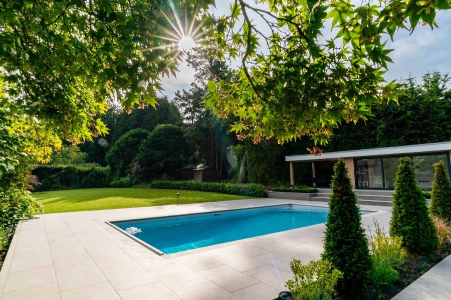 Sun beams through tree, shining light on jura grey porcelain paving used around large outdoor swimming pool, lawn and bordering hedges in background.