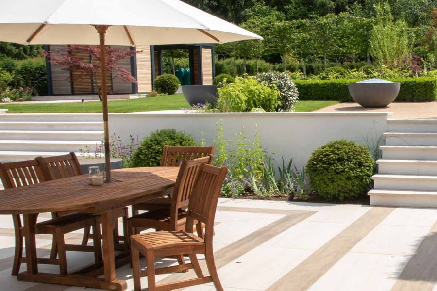 Wooden table and chair set with erected umbrella has glorious views of raised patio showing off Faro Porcelain Wall Copings.