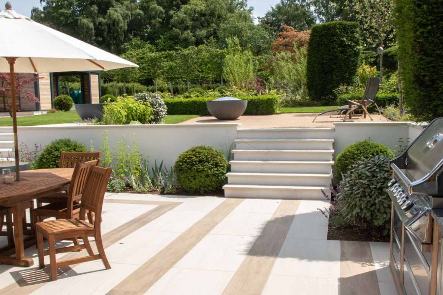 With wooden garden furniture and metal barbeque eiter side, Faro Porcelain Garden Steps lead up to lawn and water feature.