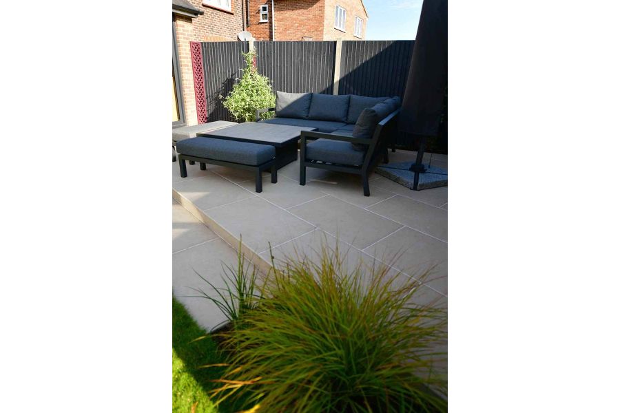 Dark outdoor furniture sits on light florence beige porcelain paving in the shade, sunlight pours over black painted fence.