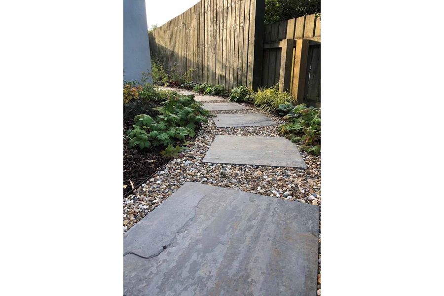 Rustic Slate Paving slabs used as stepping stones curving around path of pebbles. Flowerbeds either side.