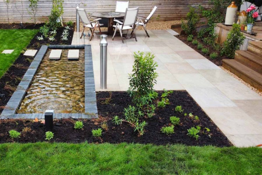 Shimmering raj green porcelain paving patio with stepping stones over brick-clad pebble pond leading to lawn.