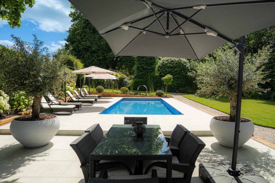 Sun shade covers glass top table with large swimming pool that uses light grey porcelain 20mm bullnose coping stones, and sunloungers in the distance.