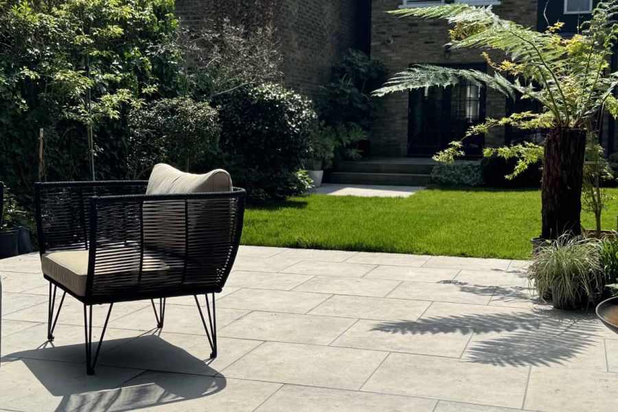 Rattan armchair sits next to small palm tree on jura grey porcelain paving, both creating shadows on these outdoor tiles.