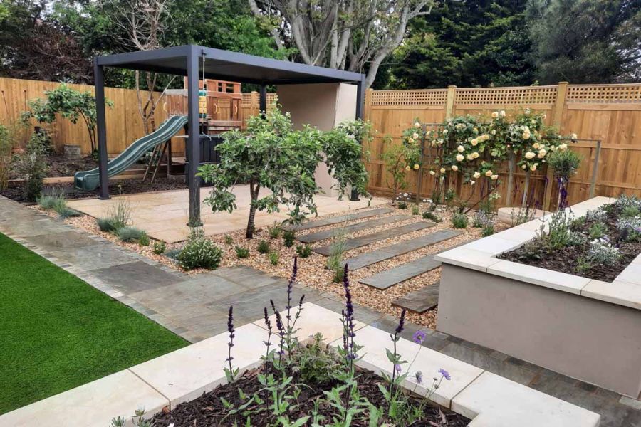 Purple plants creep out of flowerbed with harvest smooth sandstone coping stones with family garden in the background.