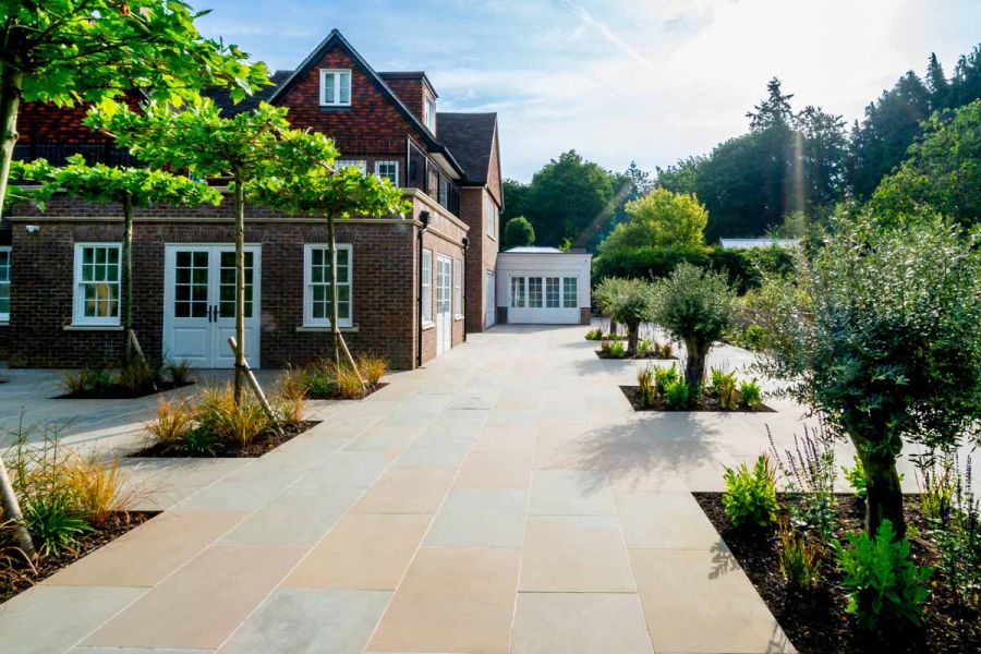 Large house sits to the left of heath sawn sandstone patio paving indented by planted trees, sunlight shows natural colour and variation.