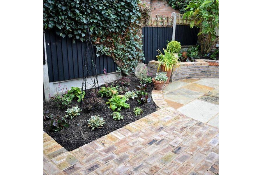 Patio of london mixture clay paver bricks flanked by flowerbed, leads to sandstone circle encased by the belgian bricks.