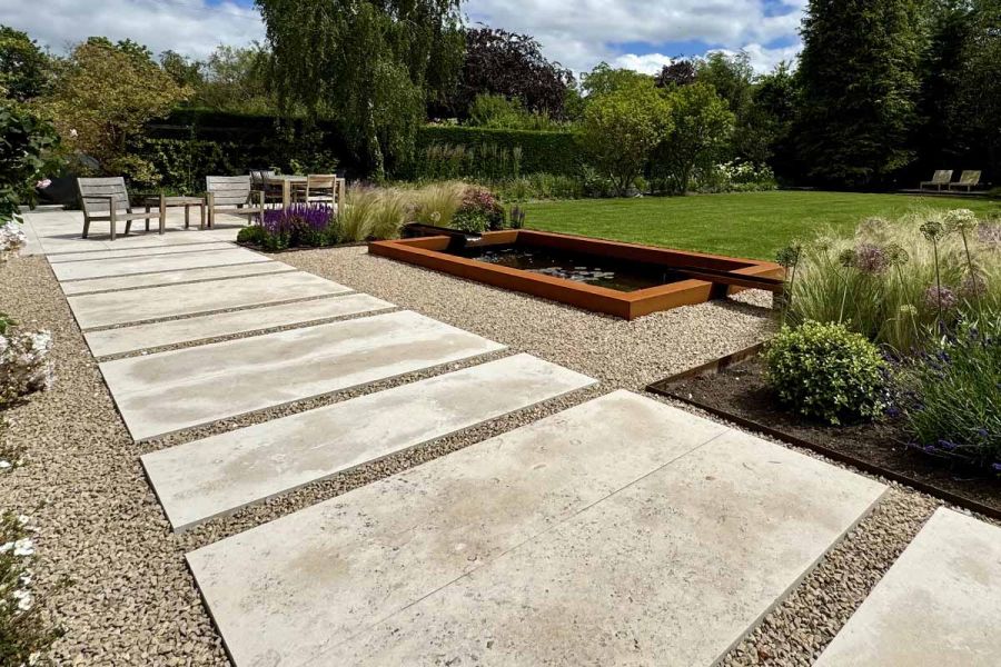 Staggered slabs of jura beige sawn limestone paving leads to patio area, walking past a corten steel framed pond.