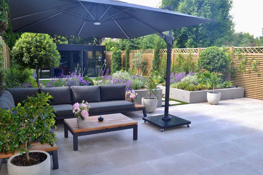 Dark sun shade covers outdoor furniture sitting on jura beige limestone paving patio with raised flowerbeds in the background.