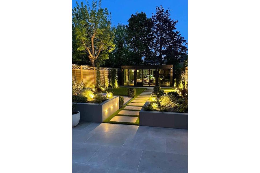 Shot at night, view down a garden shows planks of jura beige sawn limestone leading from patio to garden building.