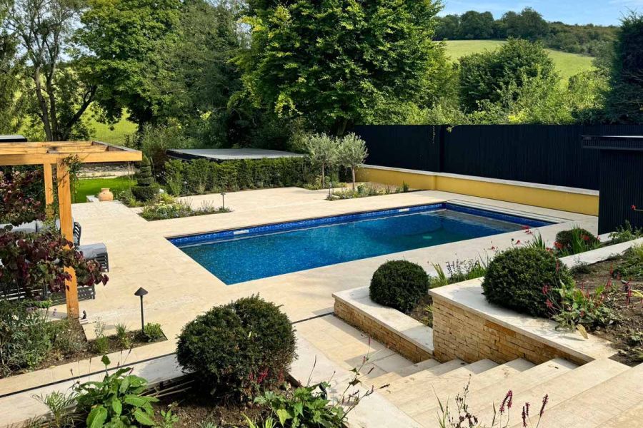 Large swimming pool surrounded by jura beige porcelain paving with matching steps in impressive garden design.