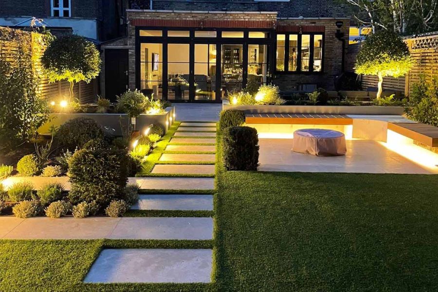 Garden lighting lights up jura beige limestone stepping stone path which leads past firepit and seating area, to garden building.