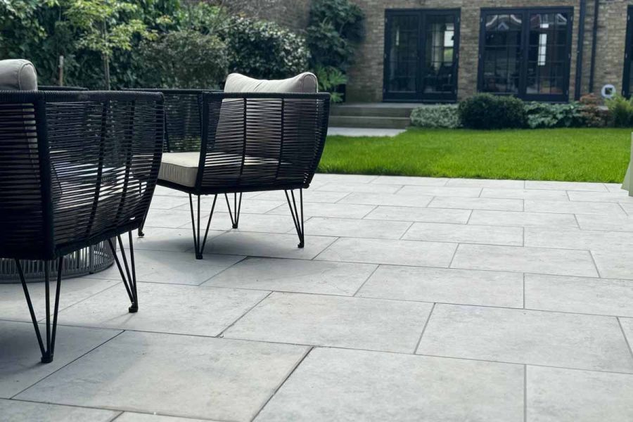 Two Rattan armchairs sit on jura grey porcelain paving, the outdoor tiles are laid next to a lawn with steps leading to house.