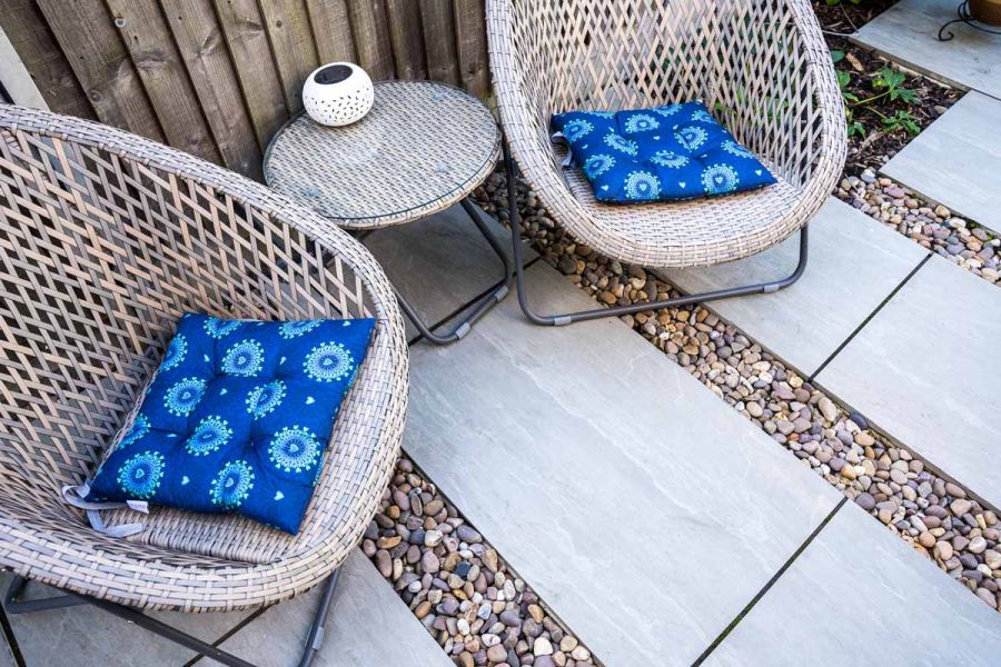 birds eye view of 2 wicker armchairs sat on kandla grey porcelain paving with pebbles indented between sets of slabs.