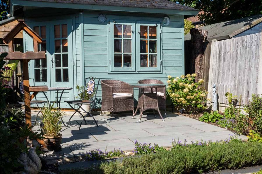 Teal coloured shed with bistro set below windows is sat on kandla grey porcelain paving in small patio setting.