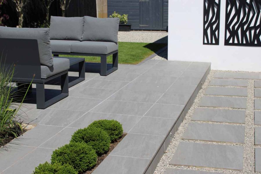 Staggered slabs in between pebbles lead to long step up to patio of kandla grey porcelain paving with modern outdoor furniture.
