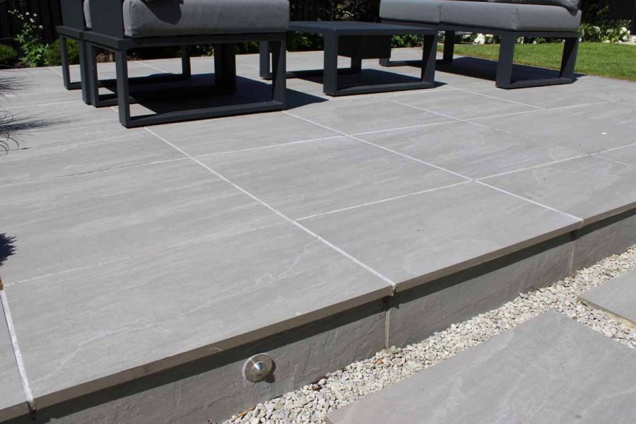 Pebble-lined staggered slabs lead up to a patio of Kandla grey porcelain paving, adorned with contemporary outdoor furniture.