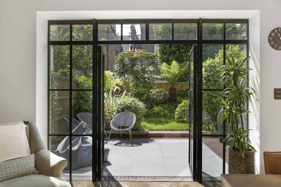 View from inside a house through frenchdoors sees a small patio with wire chairs sitting on contemporary grey sawn sandstone.